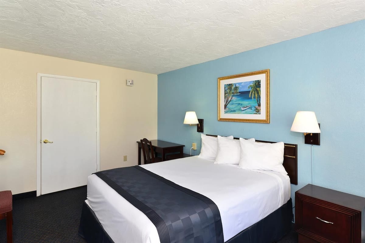 A hotel room with a bed, a desk, two lamps, a painting, and a door.