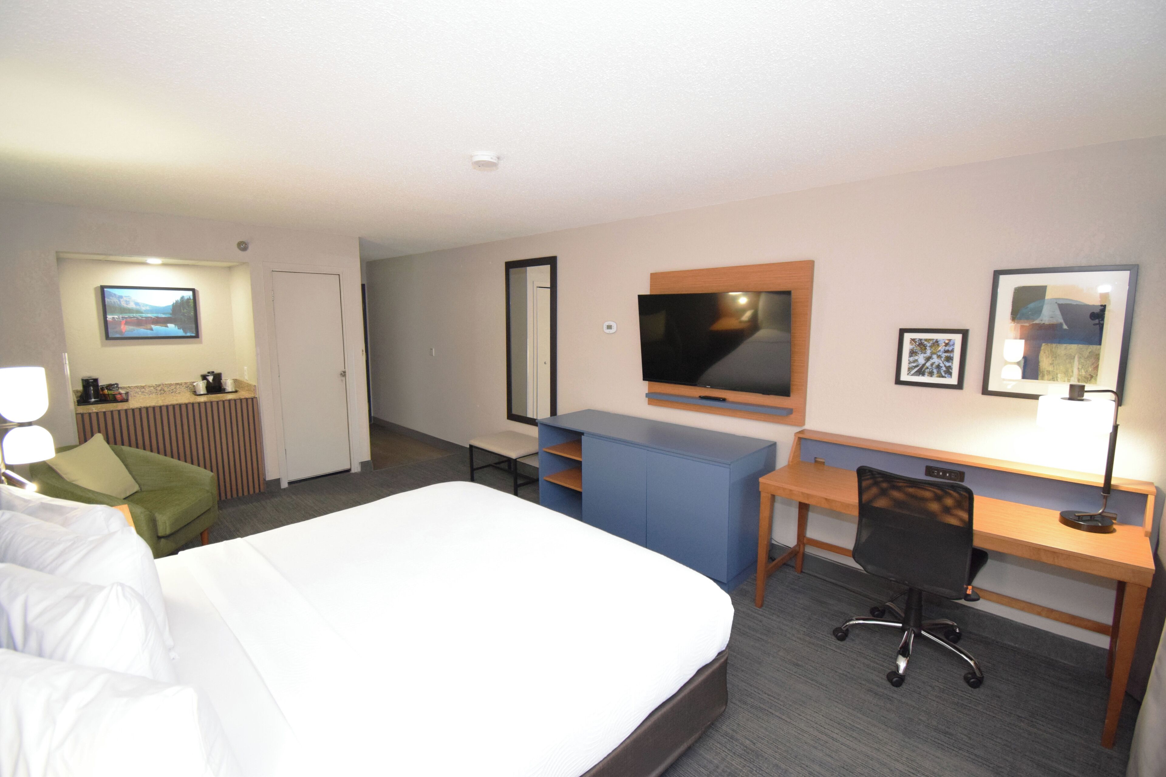 A hotel room with a bed, a desk, a chair, a TV, and a small kitchenette.