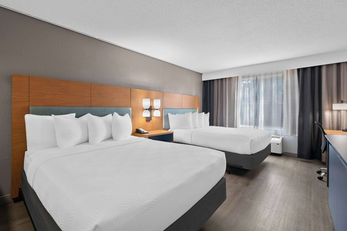 A hotel room with two beds, a wooden headboard, bedside lamps, a desk, and a window with curtains.