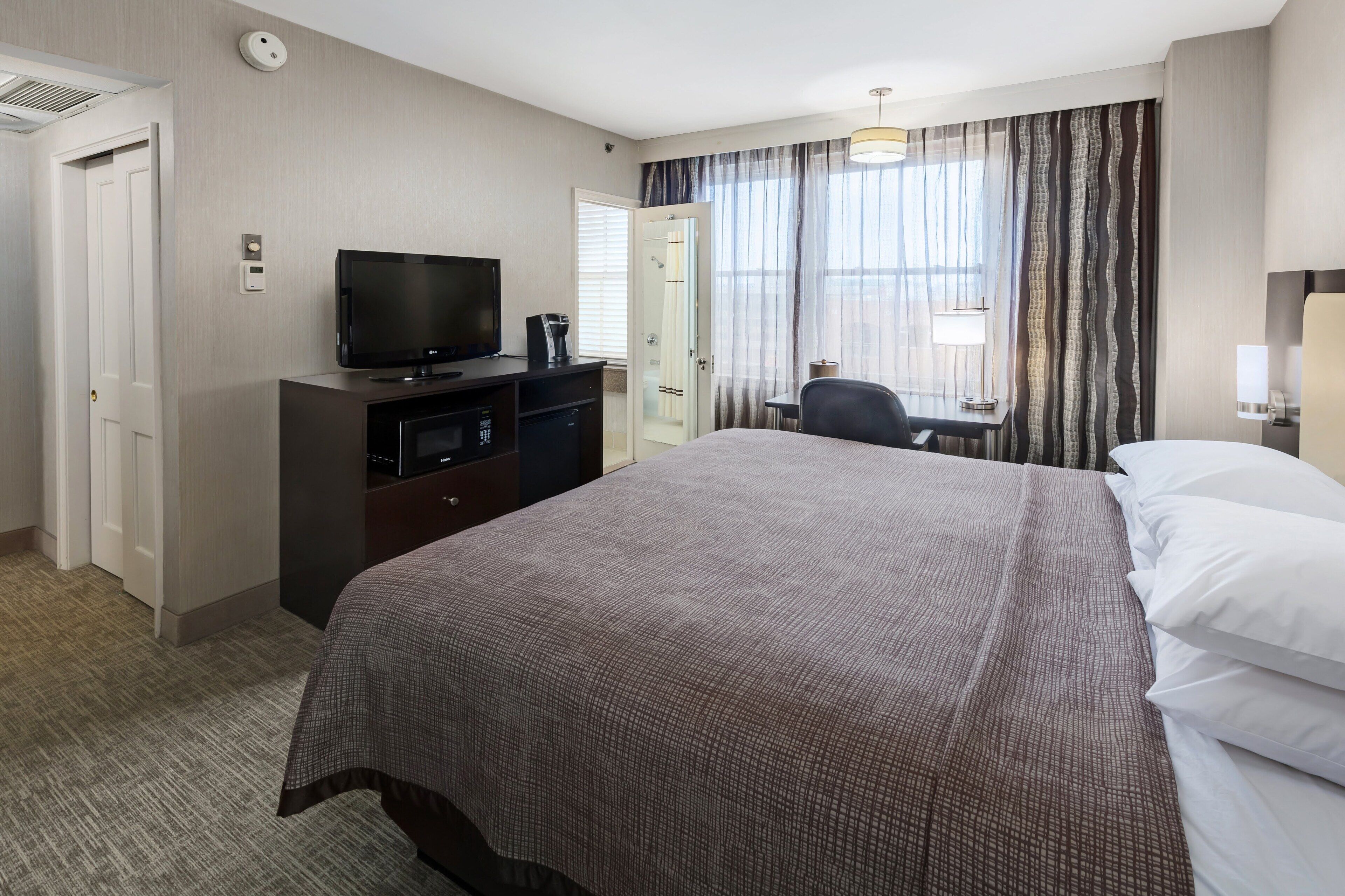 A hotel room with a large bed, a TV on a dresser, a desk with a chair, and a window with curtains.
