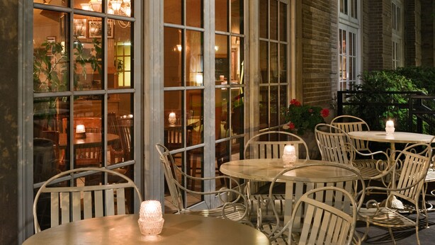 An outdoor seating area with round tables and white metal chairs, featuring candlelit tables and a view of the interior through large windows. - Park Place Hotel (Traverse City)