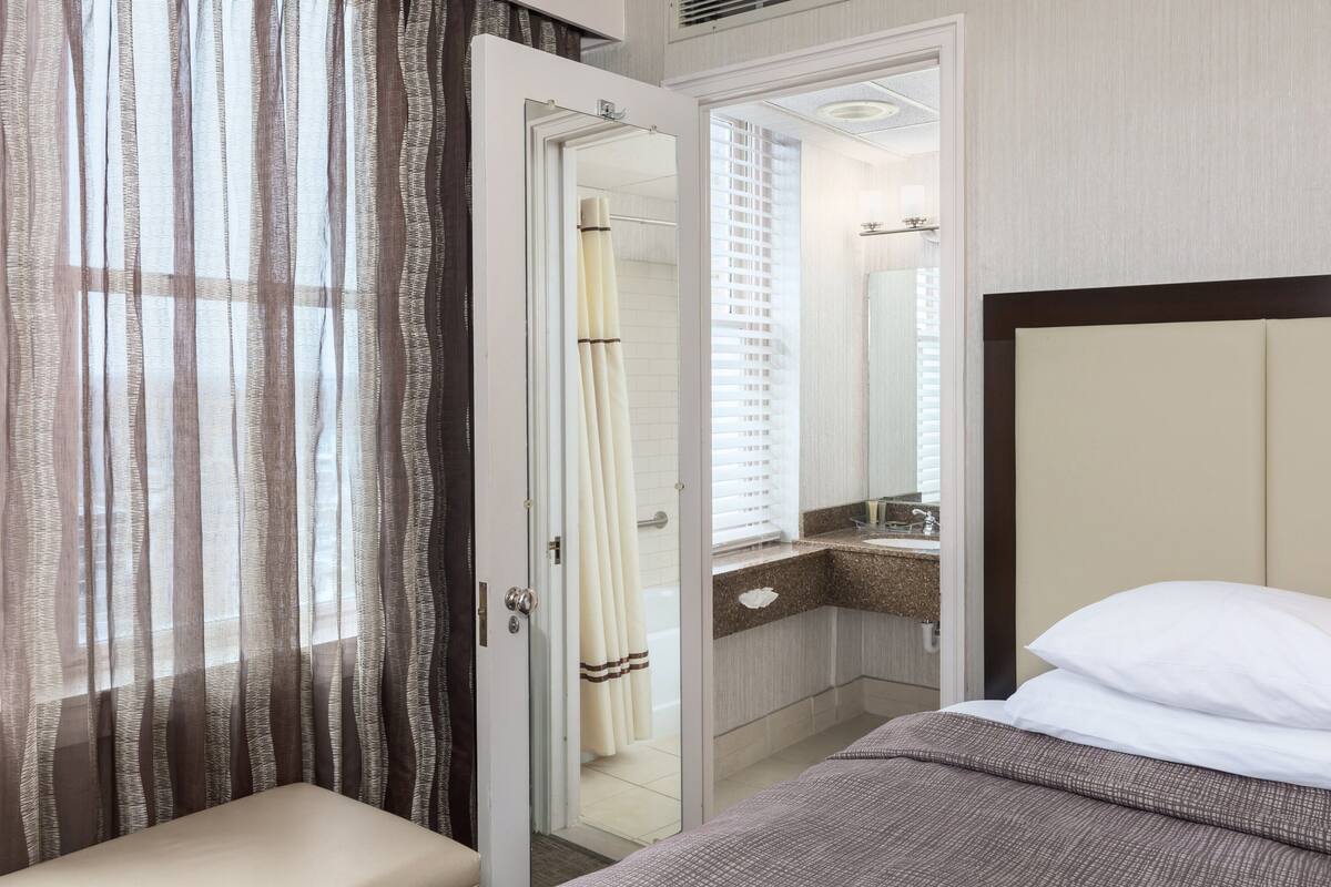 Ein Hotelzimmer mit einem Bett, einem Badezimmer mit Dusche und einem Fenster mit durchsichtigen Vorhängen.