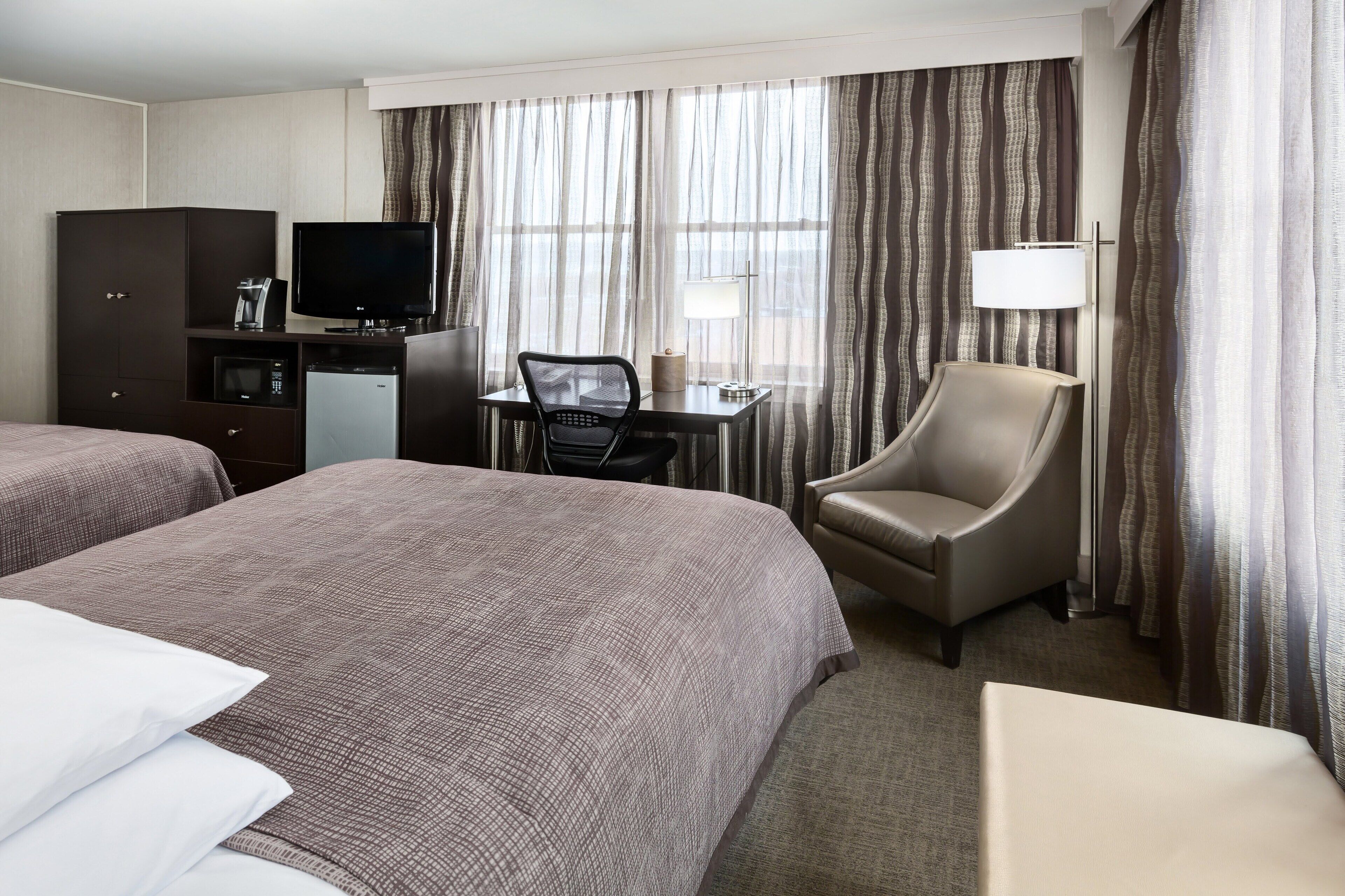 A hotel room with two beds, a desk with a chair, a television, a lamp, and a minibar.