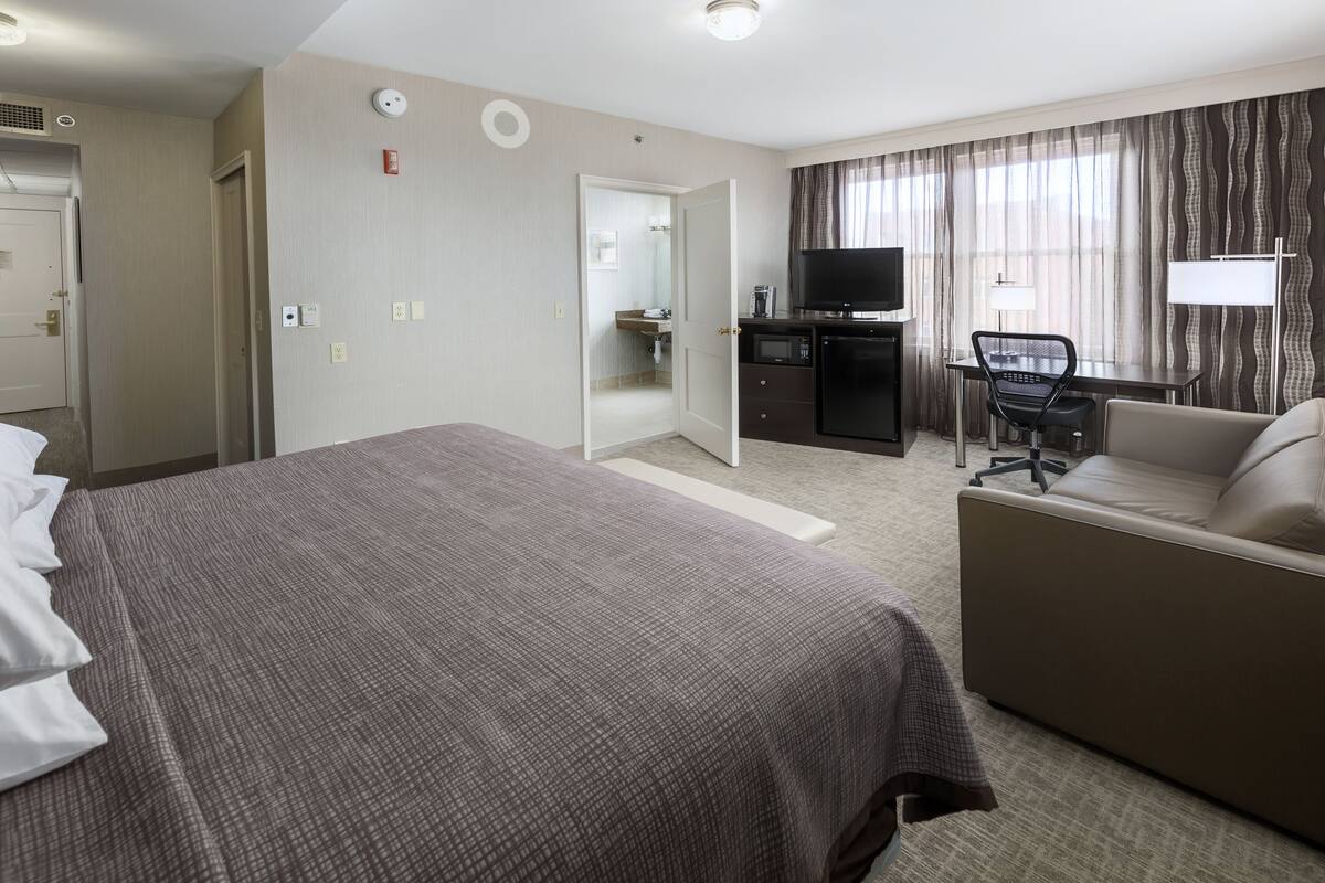 Ein Hotelzimmer mit einem großen Bett, einer Sitzecke, einem Schreibtisch mit Stuhl, einem Fernseher und einem durchgegangenen Tür erkennbaren Badezimmer.