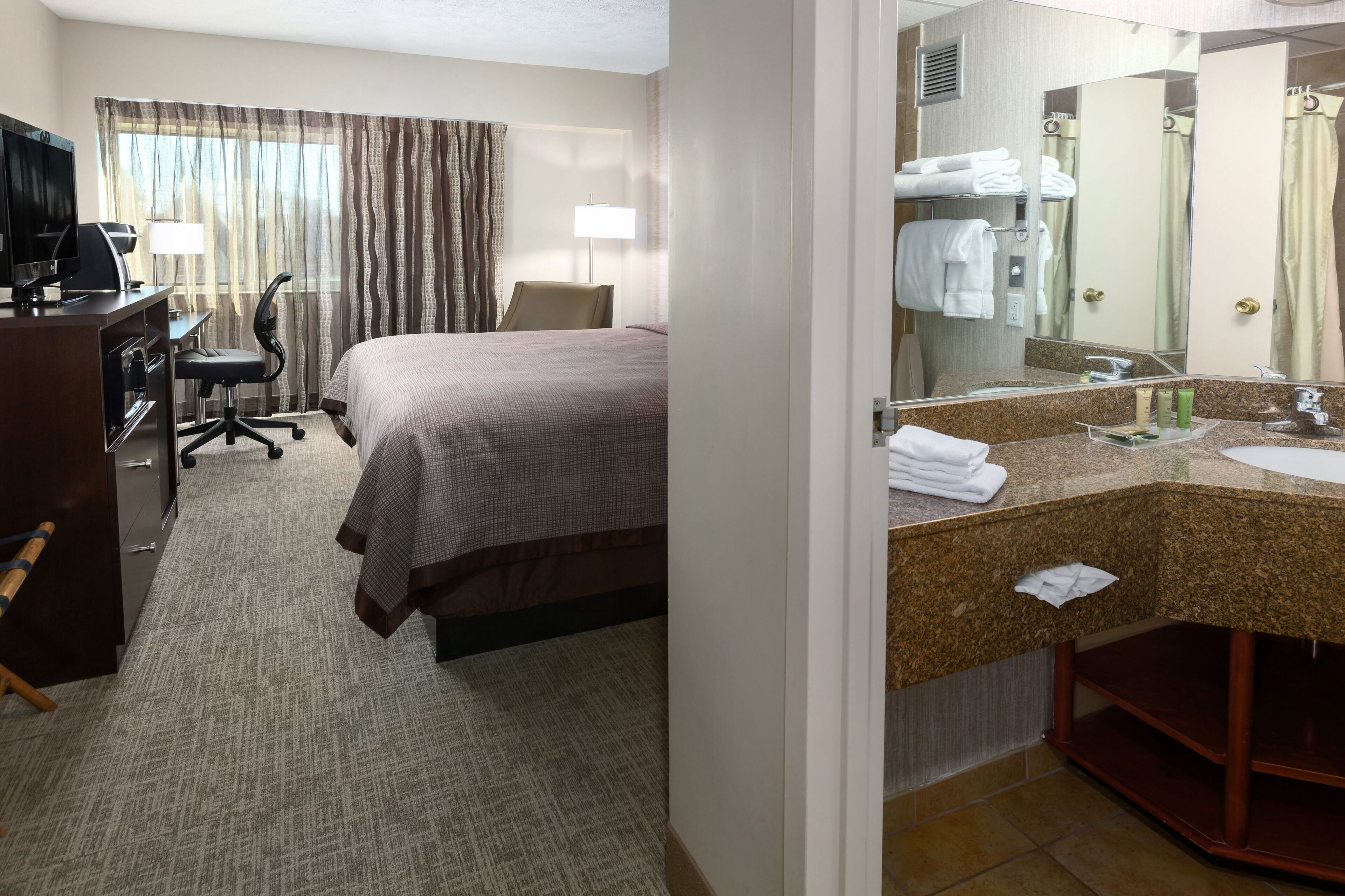 A hotel room with a bed, desk, TV, and a bathroom with a sink and mirror.