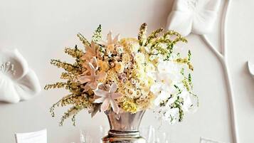 A round dining table with a white floral tablecloth, surrounded by white chairs. A large vase with a floral arrangement is placed in the center.