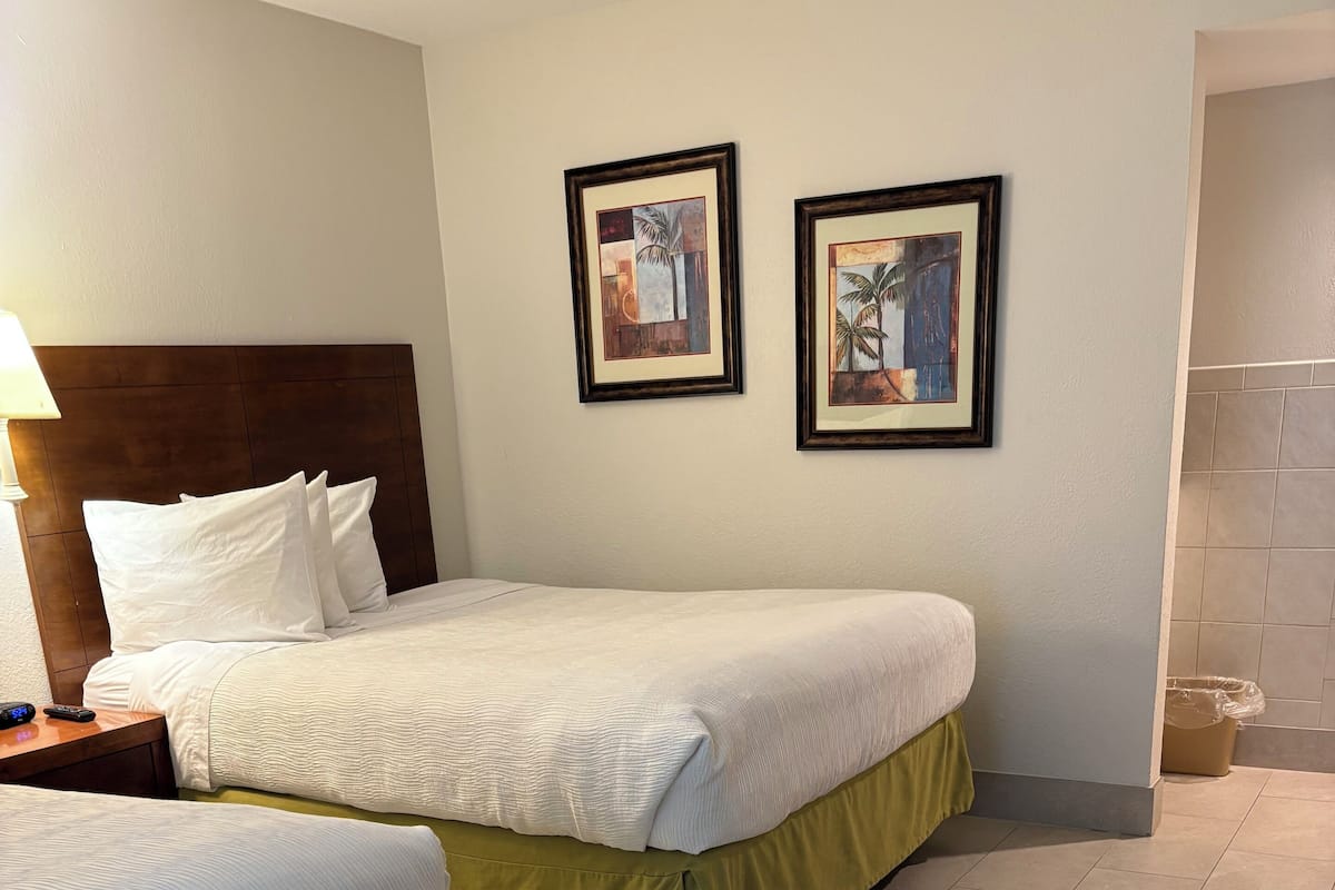 Ein Hotelzimmer mit zwei Betten, einem hölzernen Kopfteil, weißer Bettwäsche, einem grünen Bettschürze sowie einem Nachttisch mit Lampe und zwei gerahmten Bildern an der Wand.