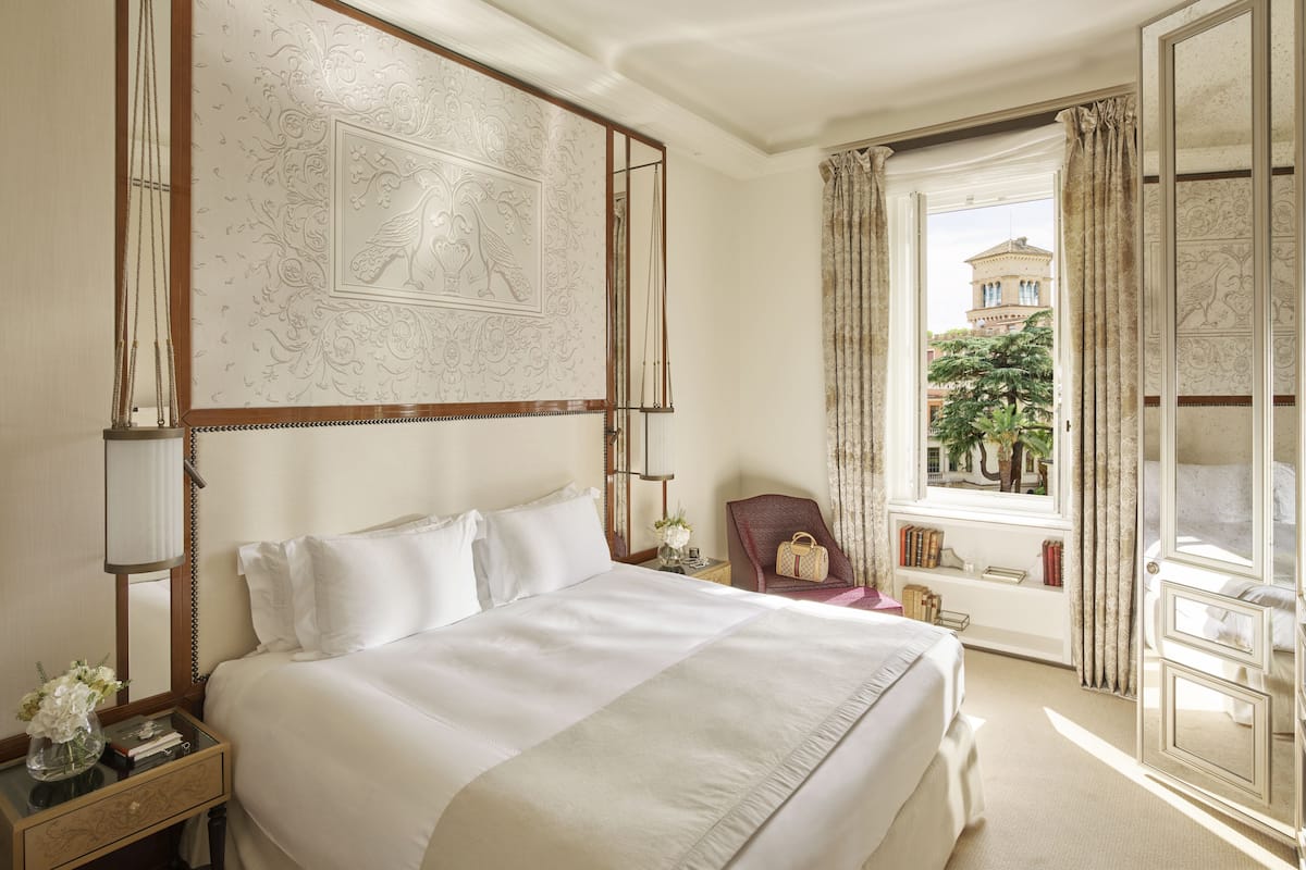 Habitación de hotel con una cama grande, una mesita de noche con lámpara, una silla, una ventana con cortinas y vistas al exterior.