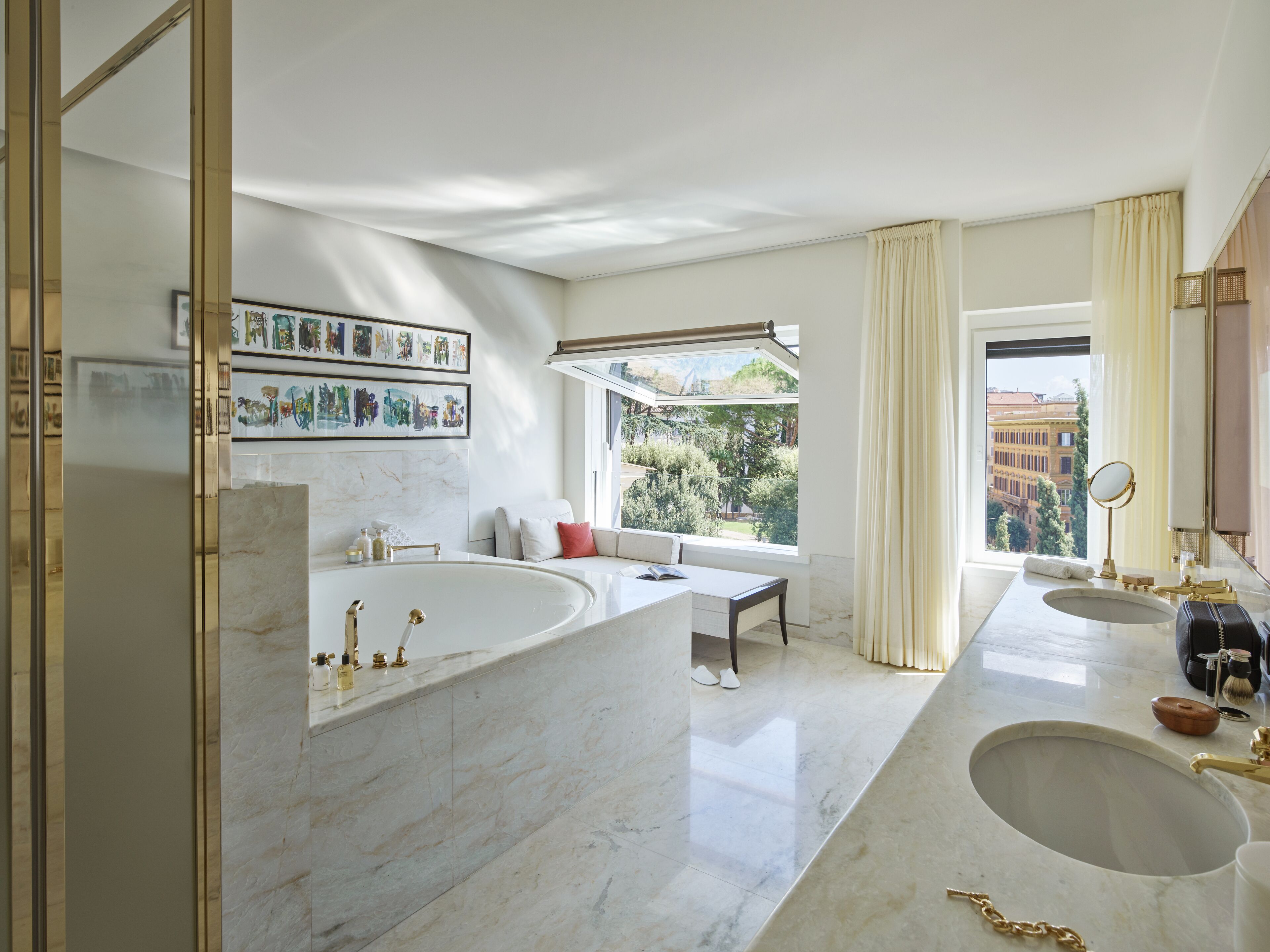 A modern bathroom with a large bathtub, two sinks, and a large window offering a view of greenery and buildings.