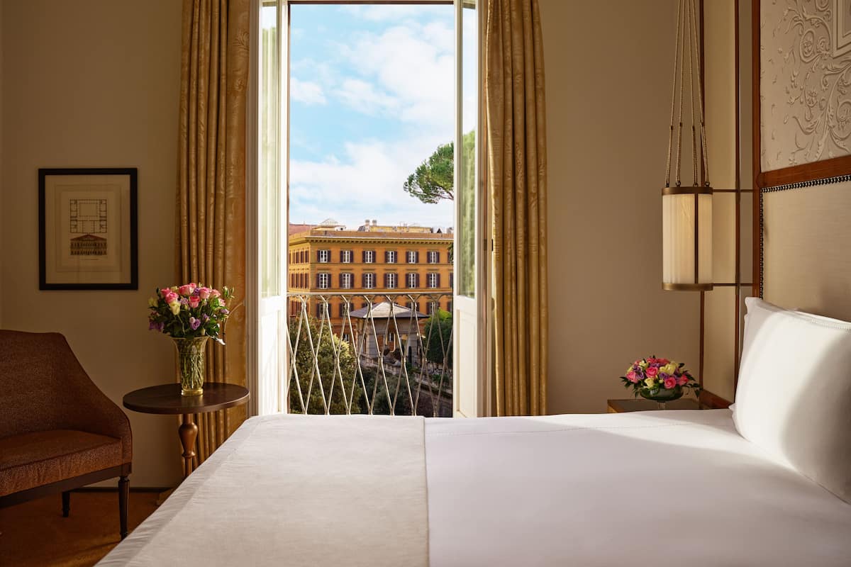 Habitación de hotel con una cama grande, una silla, una mesita con un jarrón de flores y vistas a edificios a través de una ventana con cortinas.