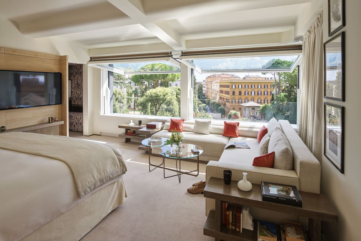 Una sala moderna con una cama grande, un sofá y una televisión. Amplias ventanas ofrecen vistas a edificios y árboles.