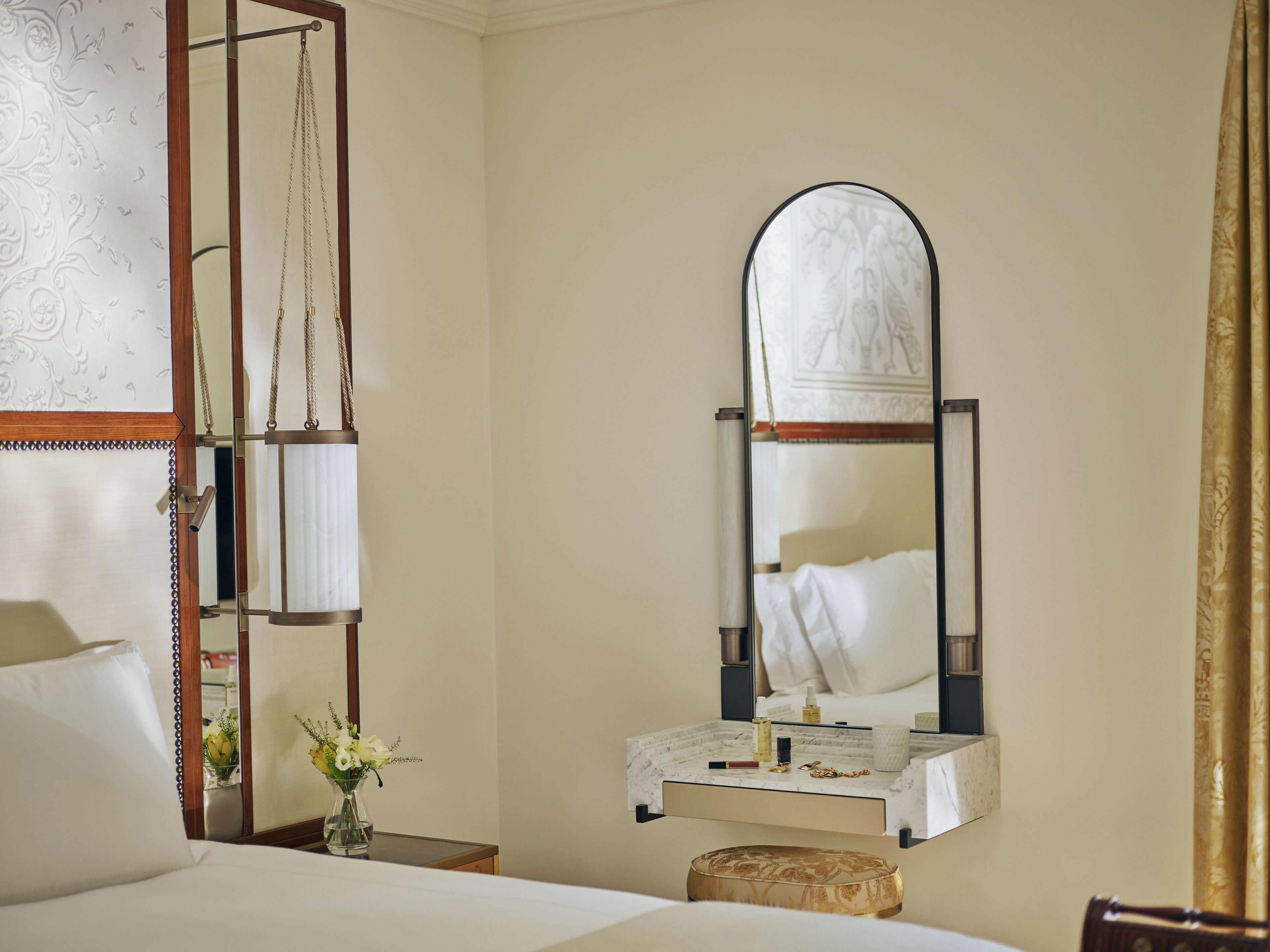 A hotel room with a bed, a vanity with a mirror, a small table with toiletries, and a vase with flowers.