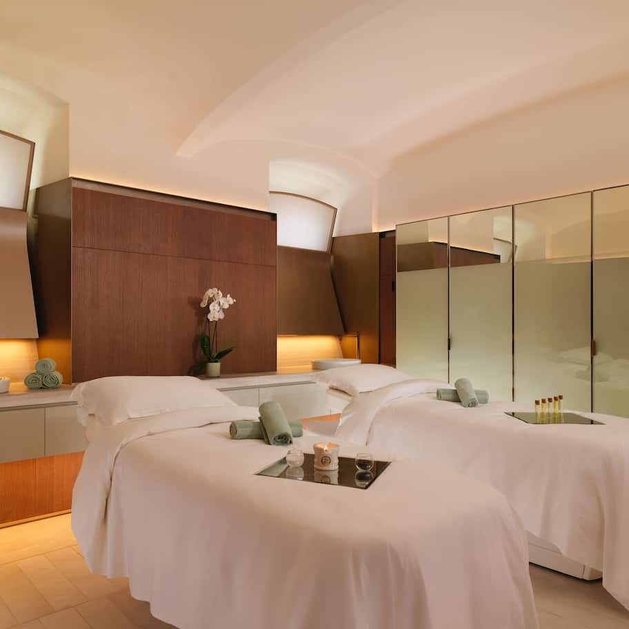 A modern hotel room with two beds, a large mirror, a bedside table with a lamp, and a glass partition wall.
