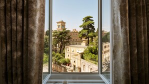 A view from a window with curtains, featuring a building with a tower and trees. - Hotel Eden - Dorchester Collection (Rome)