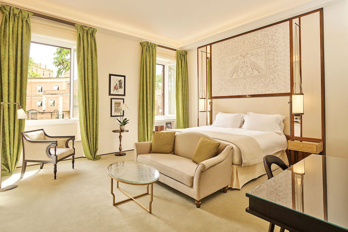 Habitación de hotel con cama, sofá, una mesita y una silla. Se ve una ventana con cortinas, una lámpara y un cuadro en la pared.