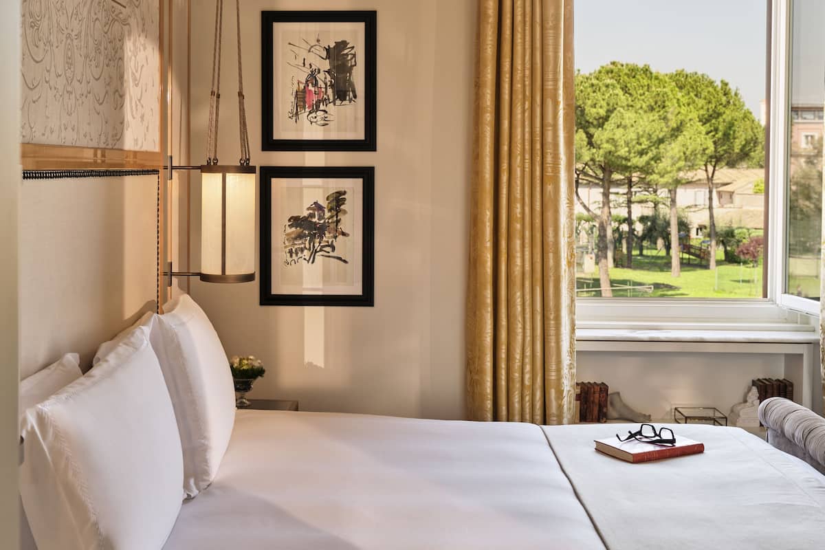Habitación de hotel con una cama grande, dos cuadros enmarcados, un ventanal con vistas a árboles y una mesita de noche con un libro y gafas.