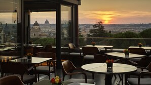 Un área de comedor al aire libre con mesas y sillas, con vistas a un paisaje urbano y un atardecer.