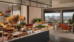A modern kitchen with a variety of fresh fruits, vegetables, and pastries. There is a view of a cityscape through large windows. - Hotel Eden - Dorchester Collection (Rome)