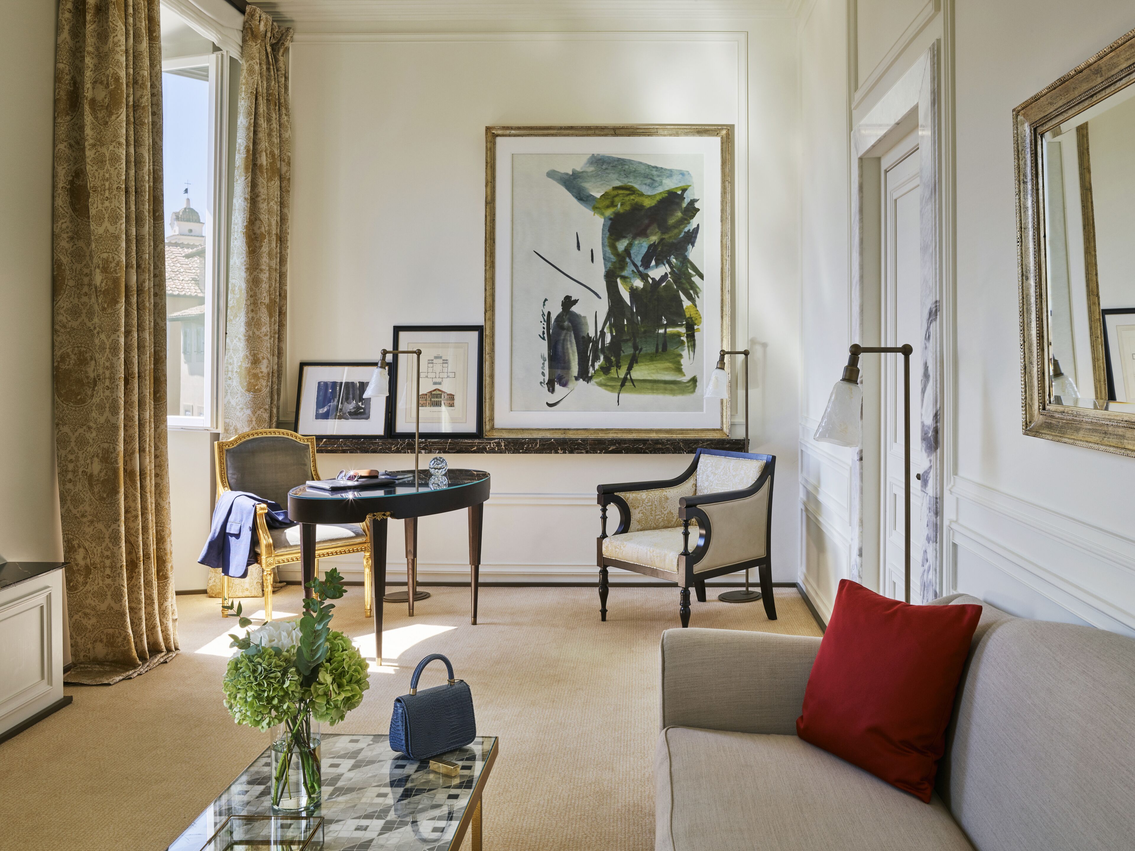 A well-lit room with a large painting, a sofa, a chair, a table with a vase, and a view of a building through a window.