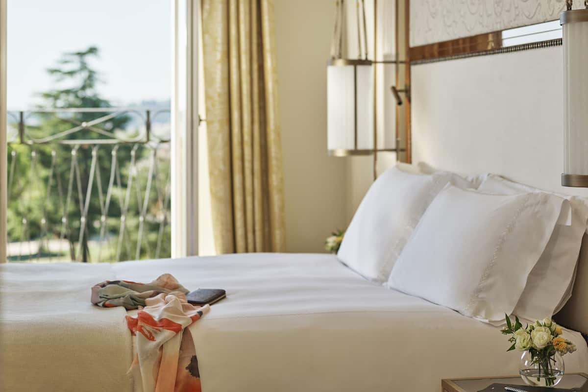 Habitación de hotel con cama, mesita de noche con un jarrón de flores, ventana con cortinas y vistas a zonas verdes.