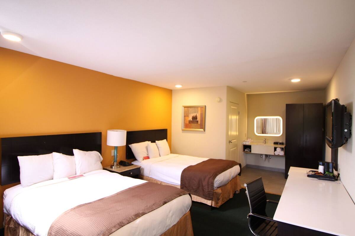 Ein Hotelzimmer mit zwei Betten, einem Schreibtisch, einem Fernseher, einem Spiegel und einem Badezimmer.