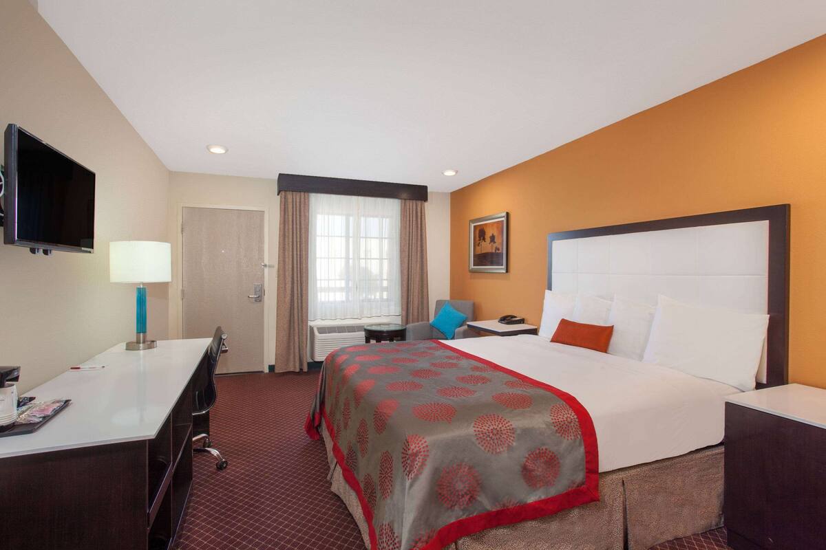 Ein Hotelzimmer mit einem großen Bett, einem Schreibtisch mit Lampe, einem an der Wand befestigten Fernseher und einem Fenster mit Vorhängen.