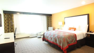 A hotel room with a large bed, a desk, a chair, a bedside table, and a window with curtains.