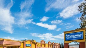 A hotel with a sign reading "RODEWAY Inn & Suites" and a parking lot with a white car. - Rodeway Inn & Suites Corona (Corona)