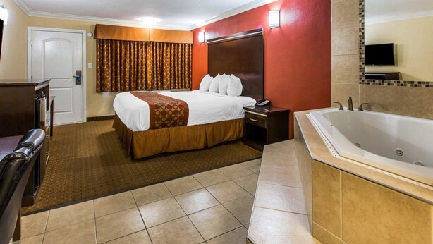 A hotel room with a bed, a desk, a chair, a TV, and a bathroom with a bathtub. - Rodeway Inn & Suites Corona (Corona)