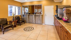 A reception area with a counter, chairs, a door, and a kitchen area with a trash bin. - Rodeway Inn & Suites Corona (Corona)
