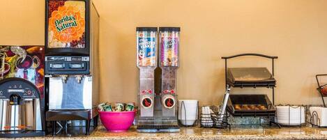 A kitchen area with a coffee machine, a fruit juice dispenser, a cereal dispenser, a grill, and a bowl of fruit.