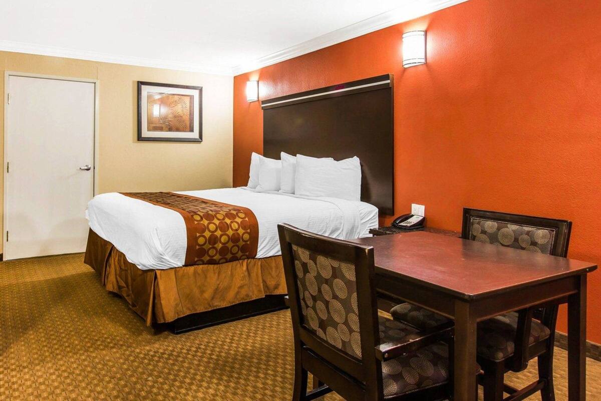 Ein Hotelzimmer mit einem Bett, einem Schreibtisch, zwei Stühlen, einem Telefon und einem gerahmten Bild an der Wand.