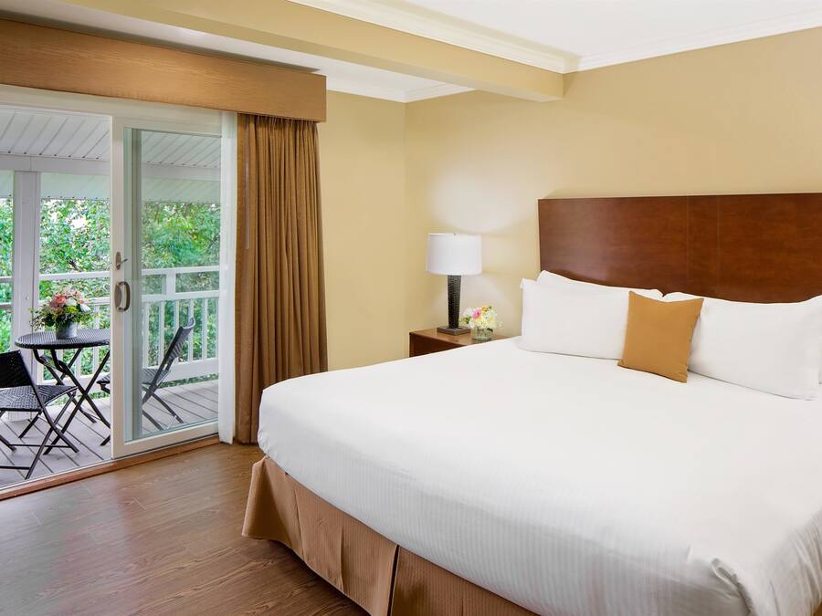 Habitación de hotel con una cama grande, una mesita de noche con lámpara, una pequeña mesa con flores y un ventanal corredero que da a un espacio exterior con sillas.