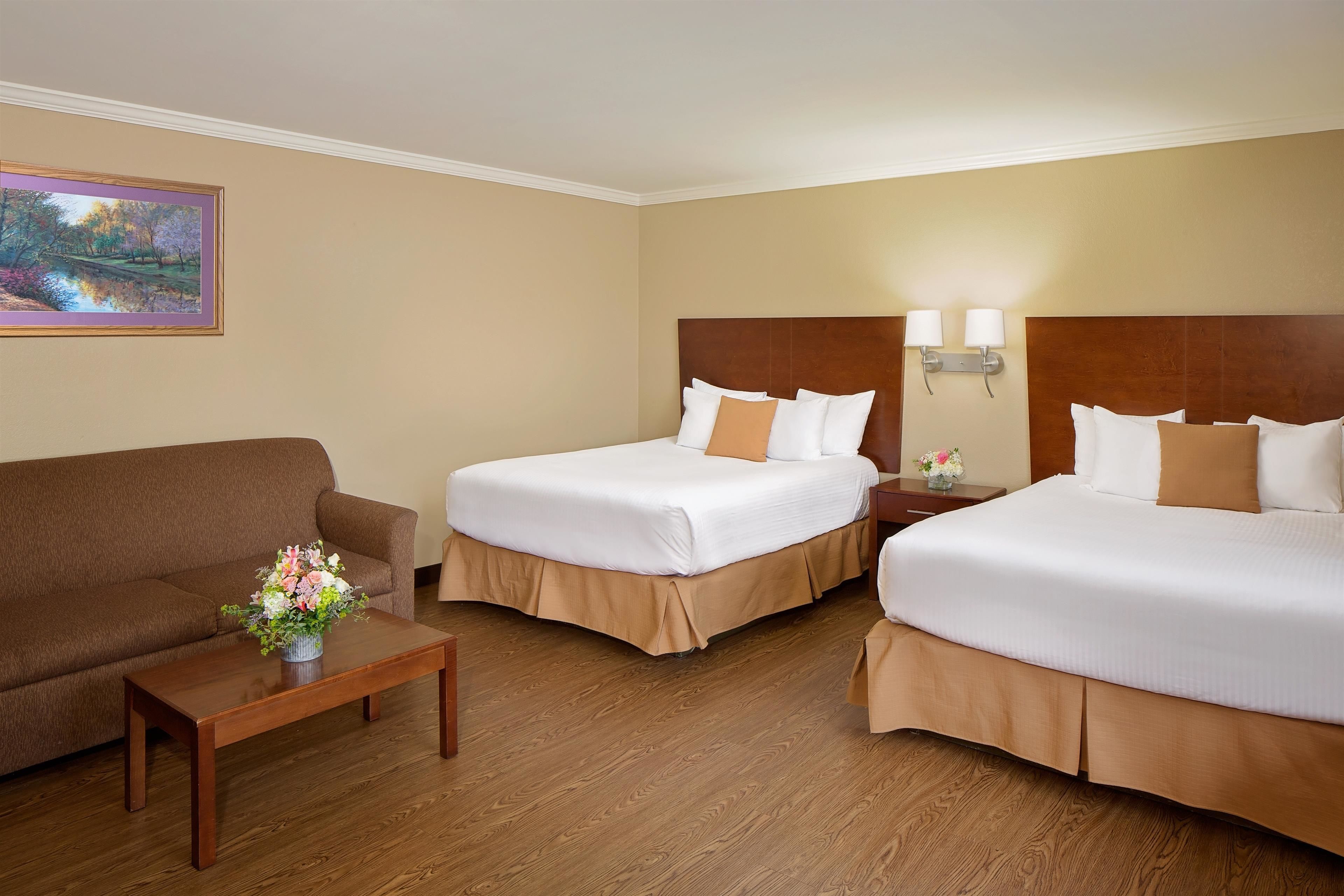 A hotel room with two beds, a sofa, a wooden floor, a painting on the wall, and a nightstand with a lamp.