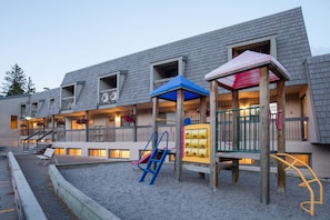 Un immeuble doté de nombreux balcons, un terrain de jeux avec un toboggan et des structures d’escalade, et un espace de repos couvert.