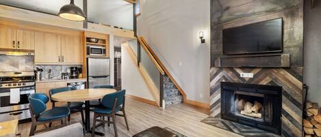 A modern kitchen with built-in appliances, a dining area with a wooden table and blue chairs, and a fireplace with a herringbone-patterned surround.