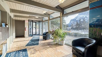 A lobby with a reception desk, a fire extinguisher, a black leather chair, a potted plant, and a view of mountains through large windows.