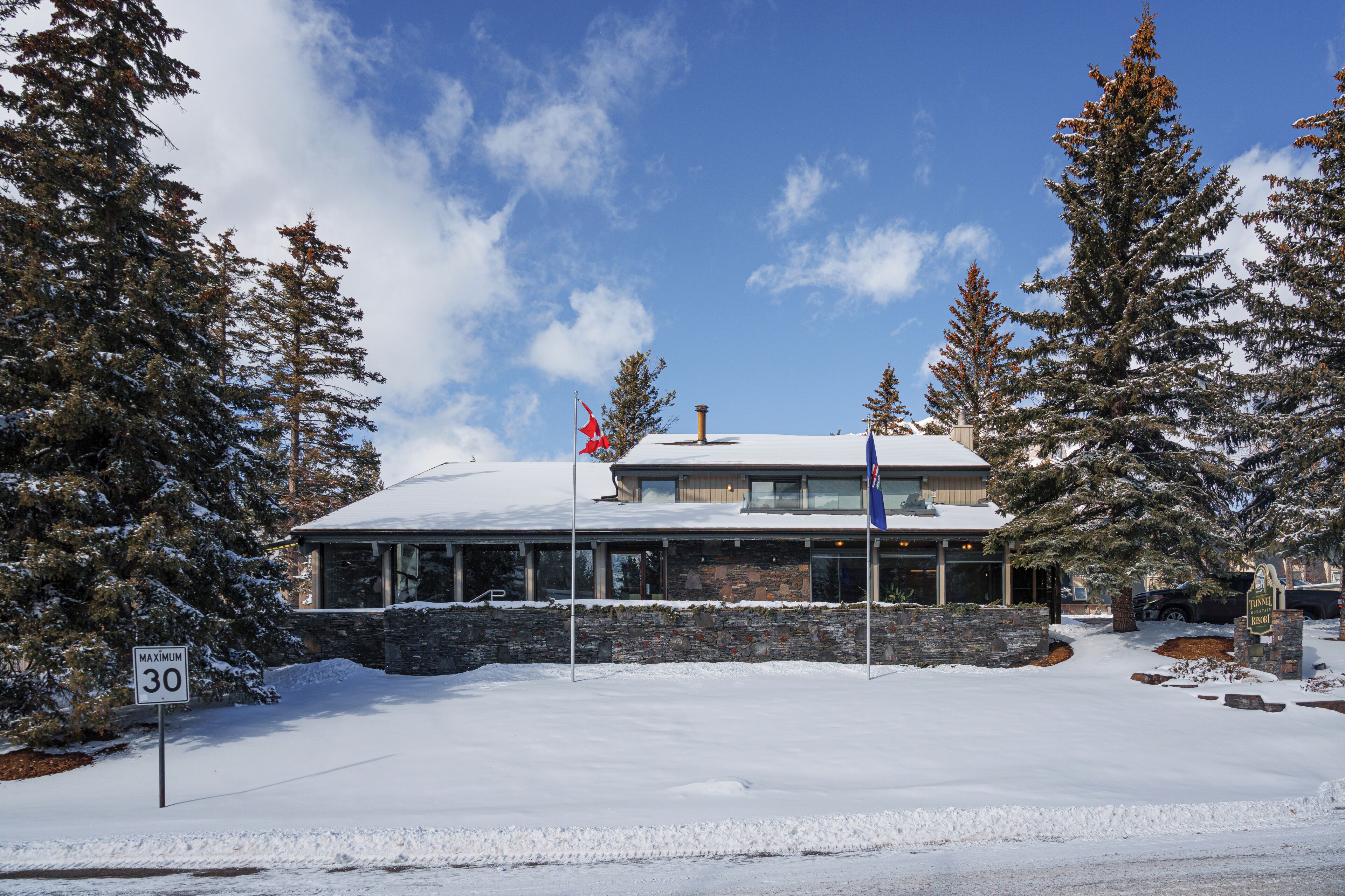 Un edificio cubierto de nieve con fachada de piedra, un asta con una bandera roja y un cartel que indica un límite de velocidad máximo de 30.