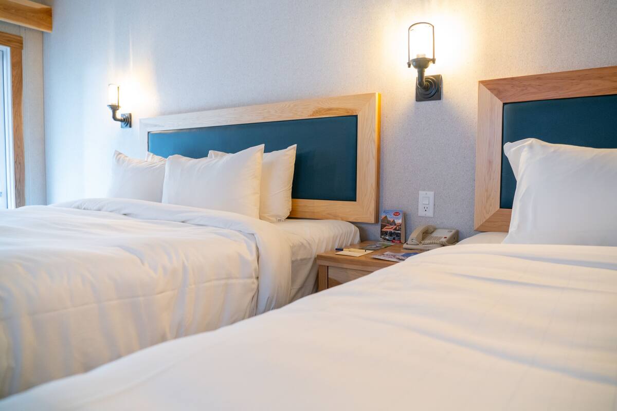 Habitación de hotel con dos camas individuales, cada una con cabecero de madera y ropa de cama blanca. Se observan mesitas de noche con lámparas y un teléfono.