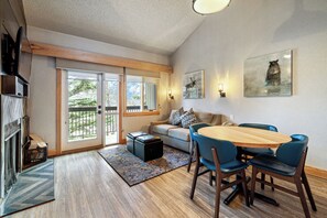 Un salon moderne avec une cheminée, une table à manger avec des chaises, un canapé et une grande fenêtre donnant sur la forêt.