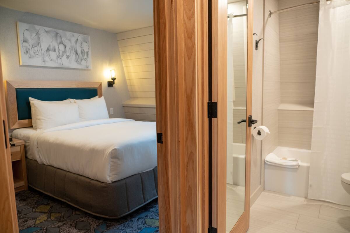Una habitación de hotel con cama, cabecero, una obra de arte colgada en la pared, mesita de noche y baño con ducha e inodoro.