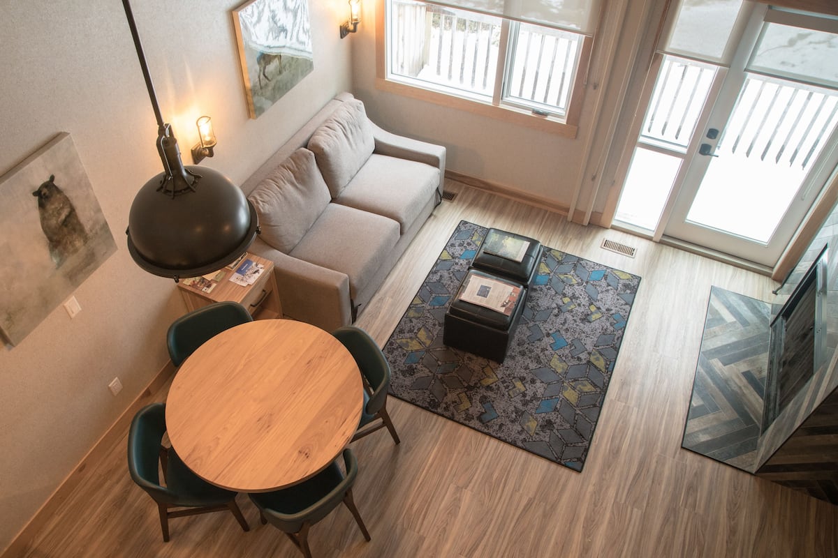 Una sala moderna con un sofá, una mesa redonda de madera y sillas. Hay una alfombra estampada en el suelo, una lámpara y obras de arte en las paredes.