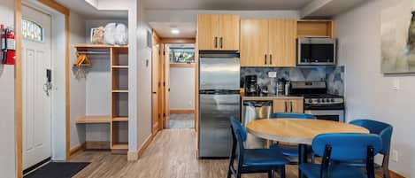 A compact kitchen with wooden cabinets, a stainless steel refrigerator, a stove, and a microwave. There is a dining table with blue chairs.