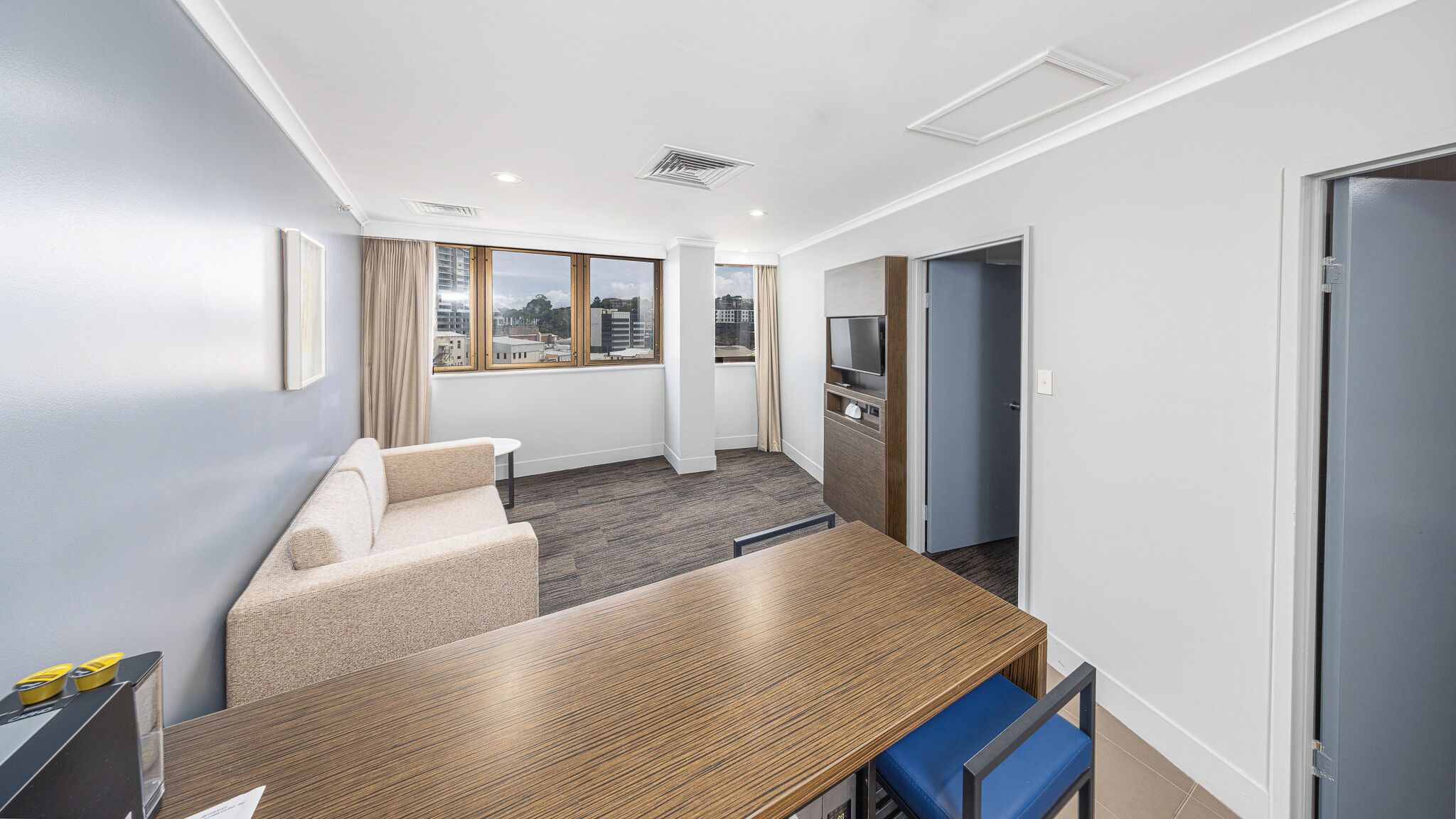 A modern hotel room with a wooden dining table, a sofa, a TV, and a view of the cityscape through large windows.
