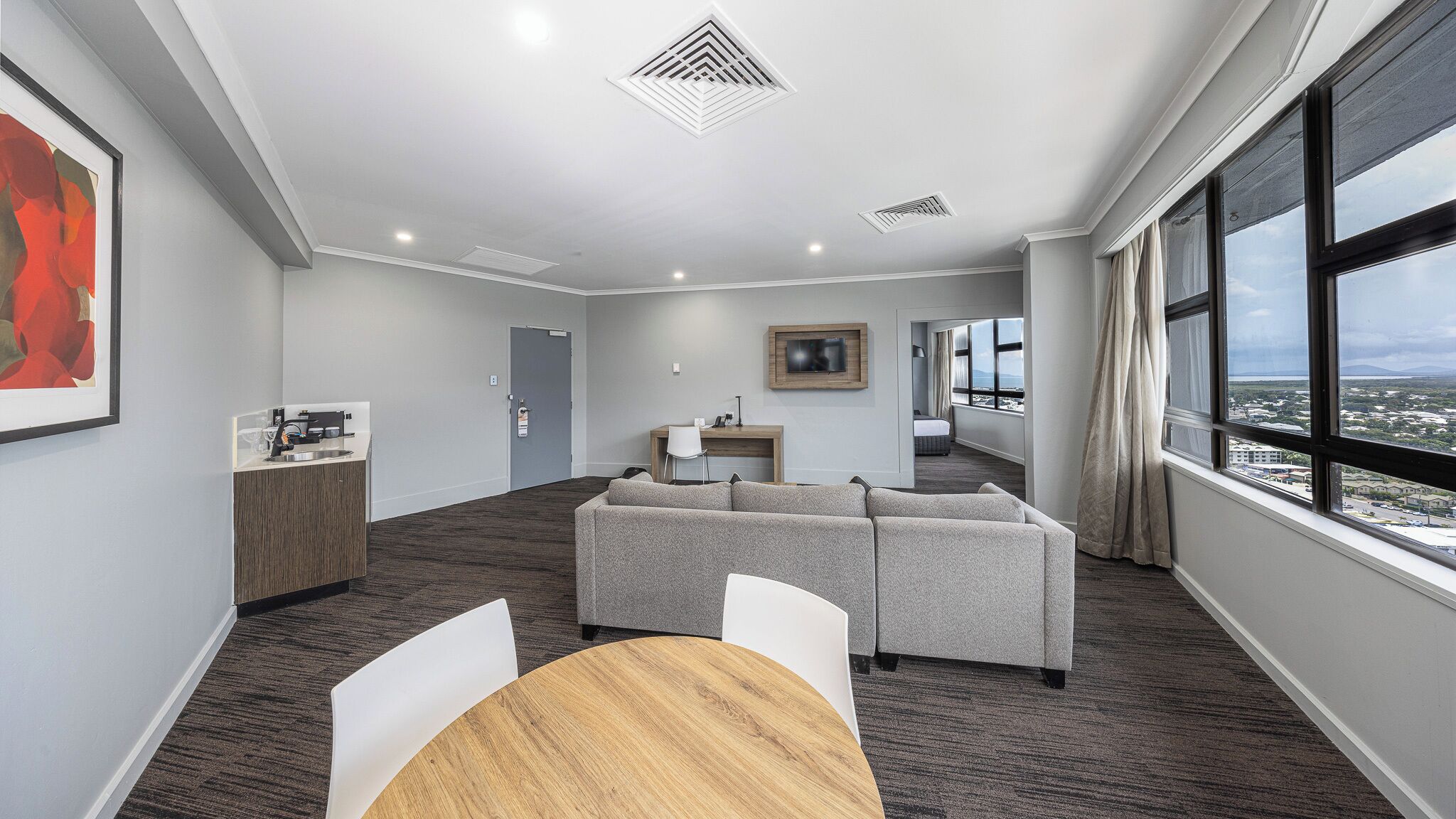 Une chambre d’hôtel moderne avec un canapé gris, une table à manger, un coin cuisine et de grandes fenêtres donnant sur la ville.