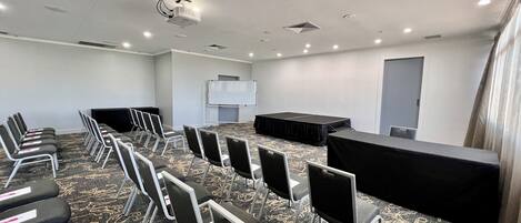 A conference room with rows of chairs, a stage with a black cloth, a whiteboard, and a projector on the ceiling.