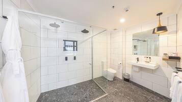 Um banheiro moderno com chuveiro em gabinete de vidro, azulejos brancos, piso de mármore, vaso sanitário e toucador com espelho e prateleiras.
