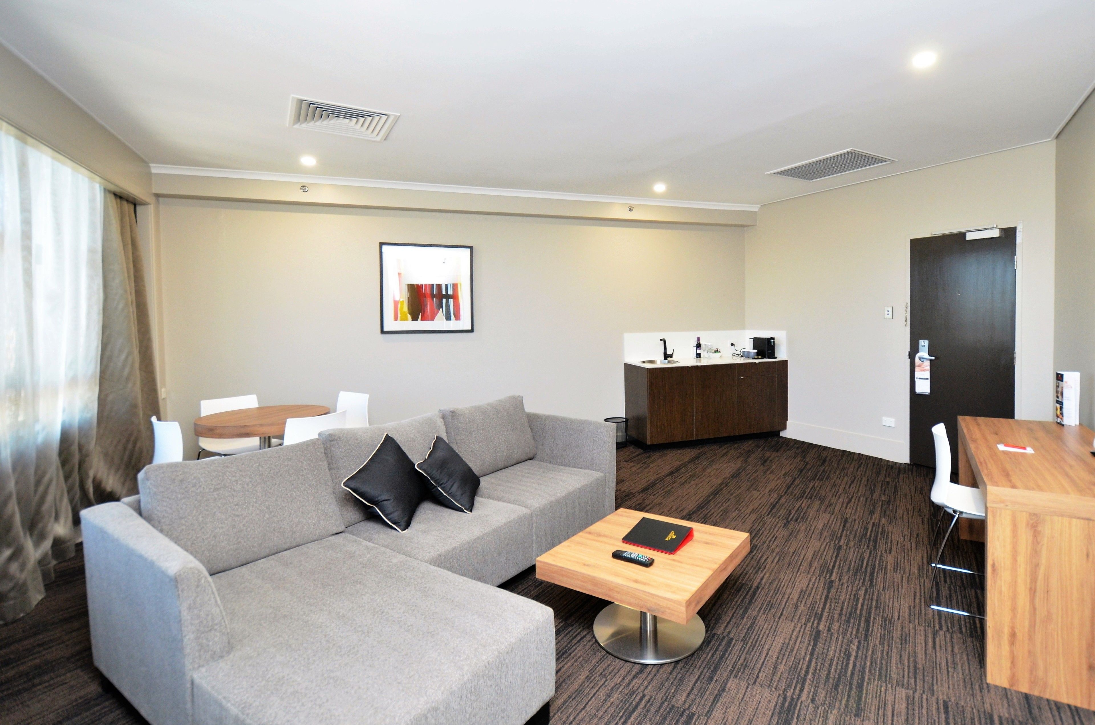 Una habitación de hotel moderna con un sofá gris, una mesa de centro de madera, una mesita redonda y una zona de kitchenette.