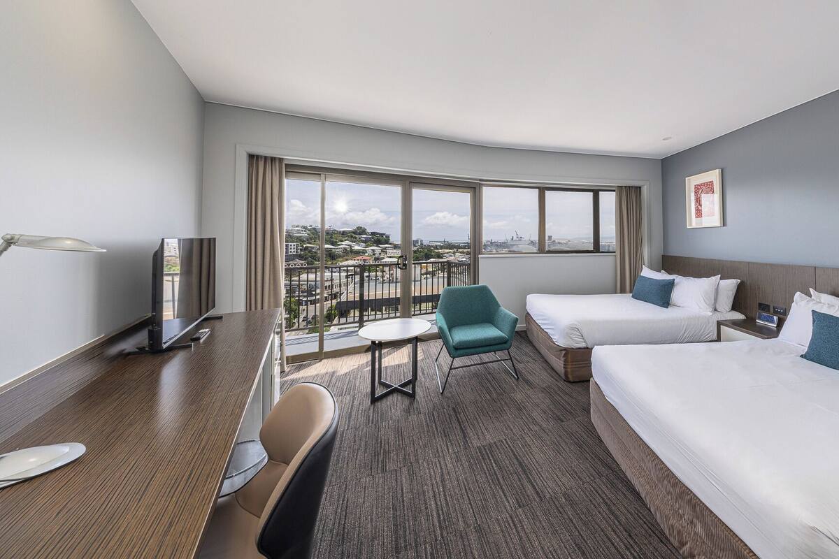 Une chambre d’hôtel avec deux lits, un bureau, une chaise, une télévision et un balcon donnant sur la ville.