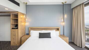 A hotel room with a large bed, wooden headboard, bedside tables, a wardrobe, and a view of the city through the window.